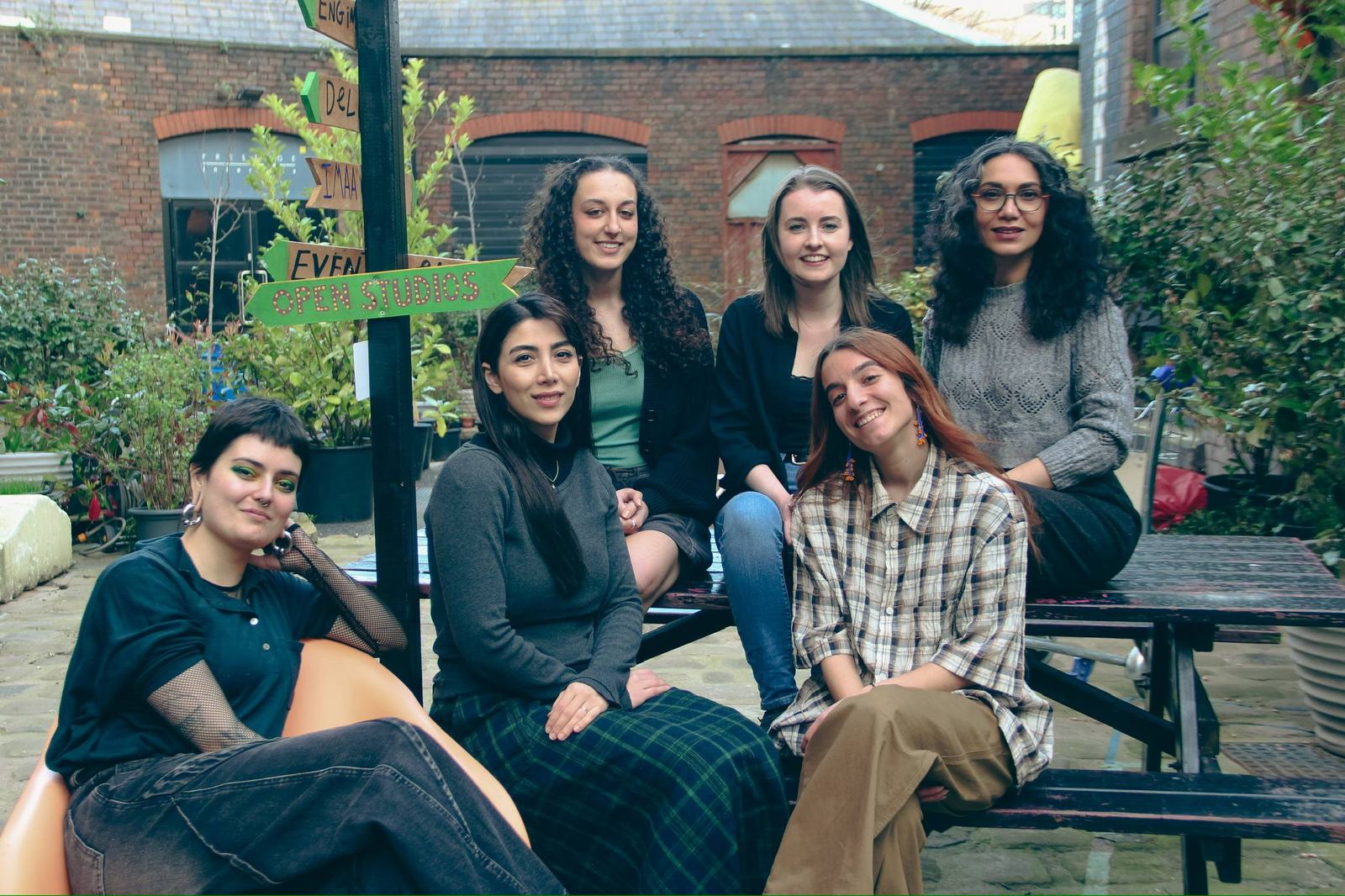 Six members of 1838 Collective sitting together in a courtyard in Manchester, beside a hand-painted sign that reads “Open Studios.”